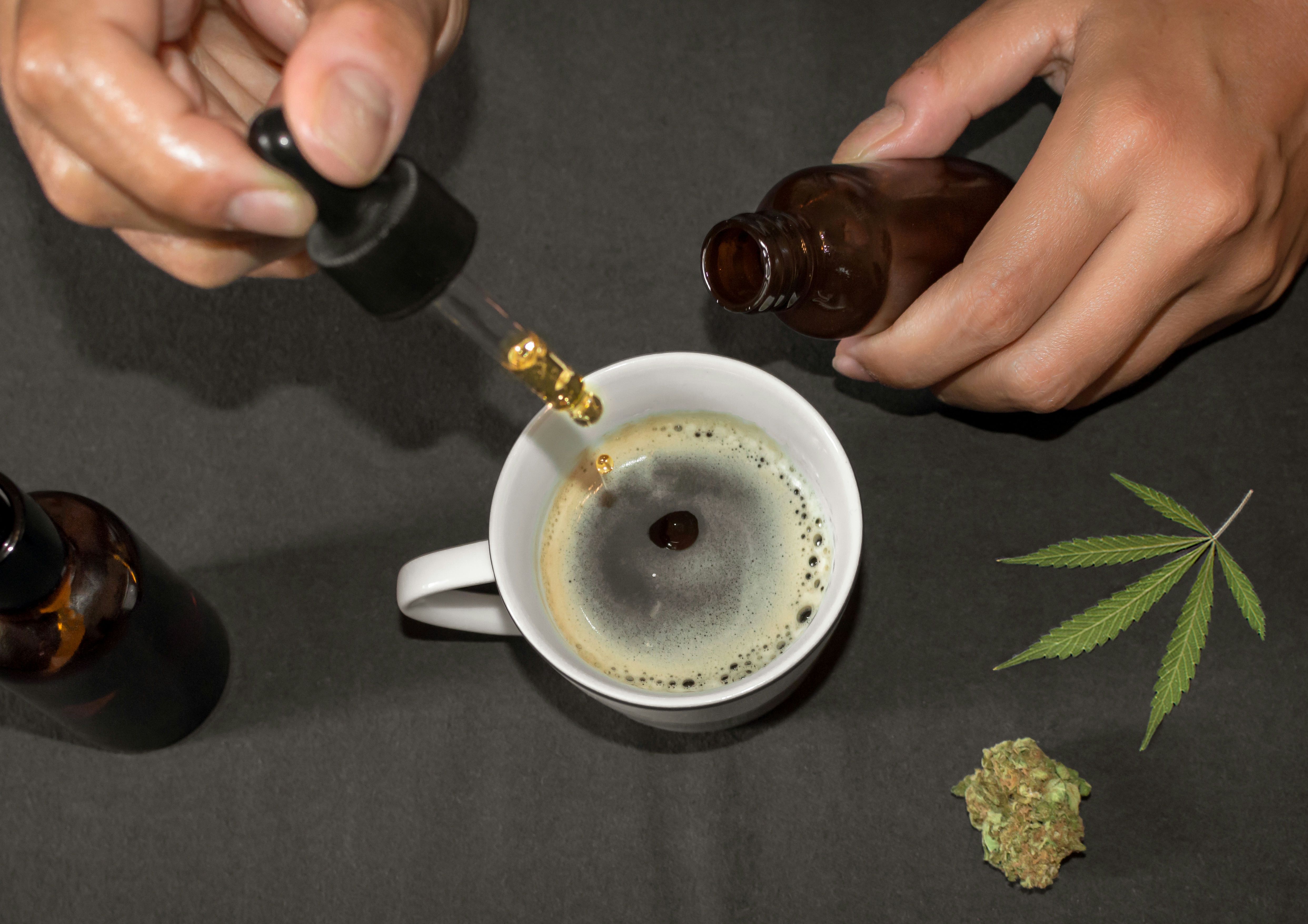 Pouring CBD tincture in a coffee cup, natural remedy of marihuana. Person using cannabis oil with a dropper in a table with marihuana leaf.