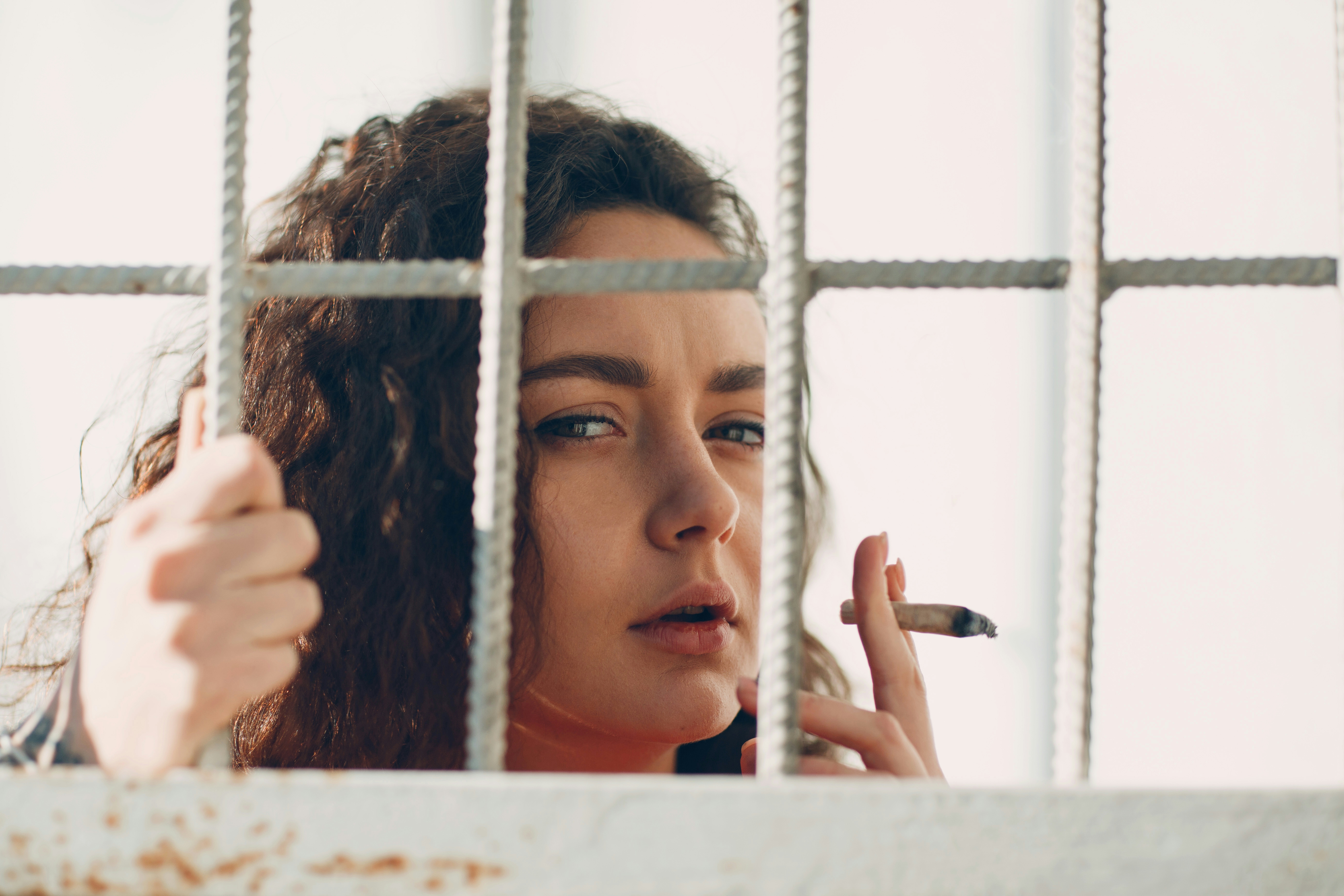 Young brunette curly woman in orange suit behind jail bars smokes marijuana joint. Female in colorful overalls portrait.