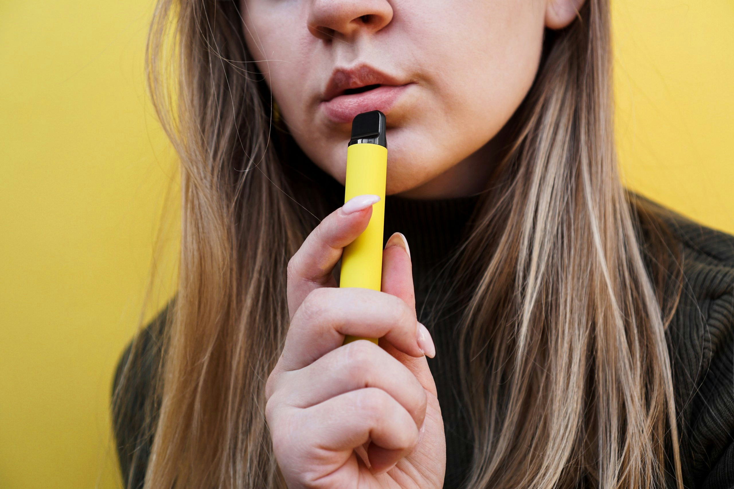 Close up of a young girl smokes a disposable electronic cigarette. Bright yellow background. Alternative to regular cigarettes and vape