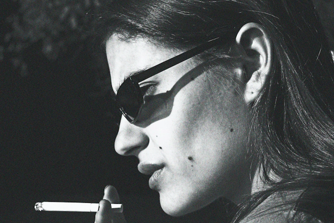 A side profile of a woman with black sunglasses on that has a joint close to her mouth.