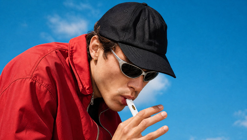 Person is inhaling from a disposable vape, wearing a bright red jacket, black cap, sunglasses, and has a bright sky background behind them.