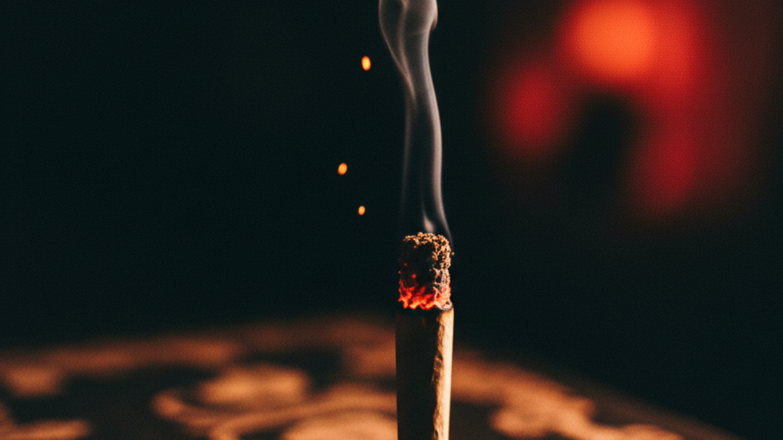 Close-up of a lit joint ember glowing under red party light with smoke curling into darkness
