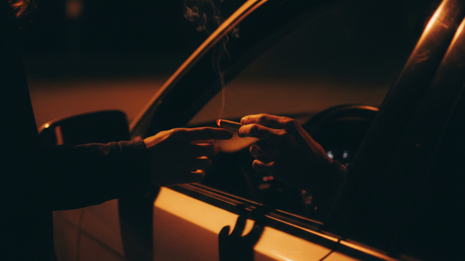 Tight crop of two hands passing a lit joint through a car window at night, illuminated by dashboard glow and ember light