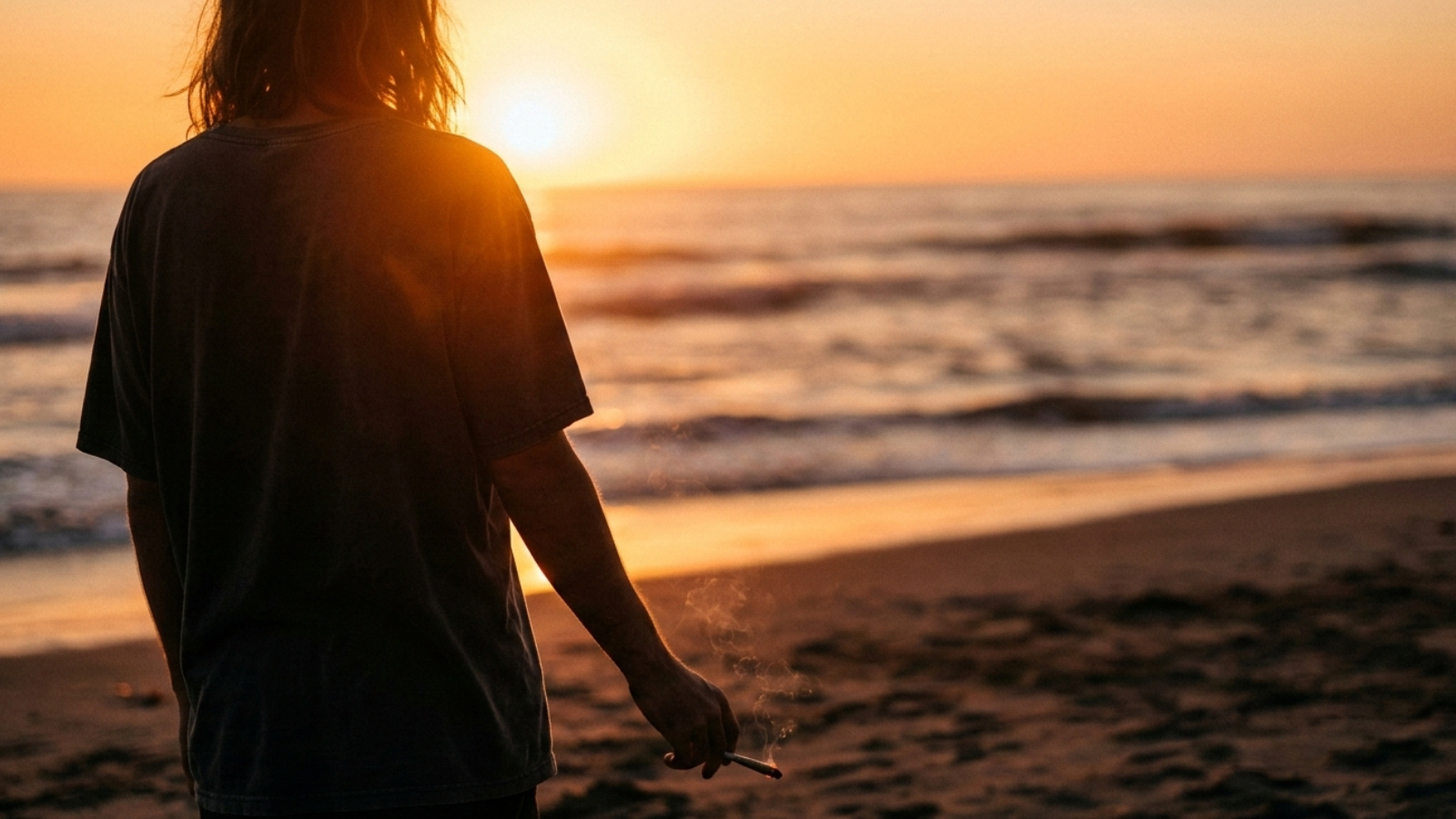 Over-shoulder view of a person holding a joint while watching the sun set over the ocean in soft golden light