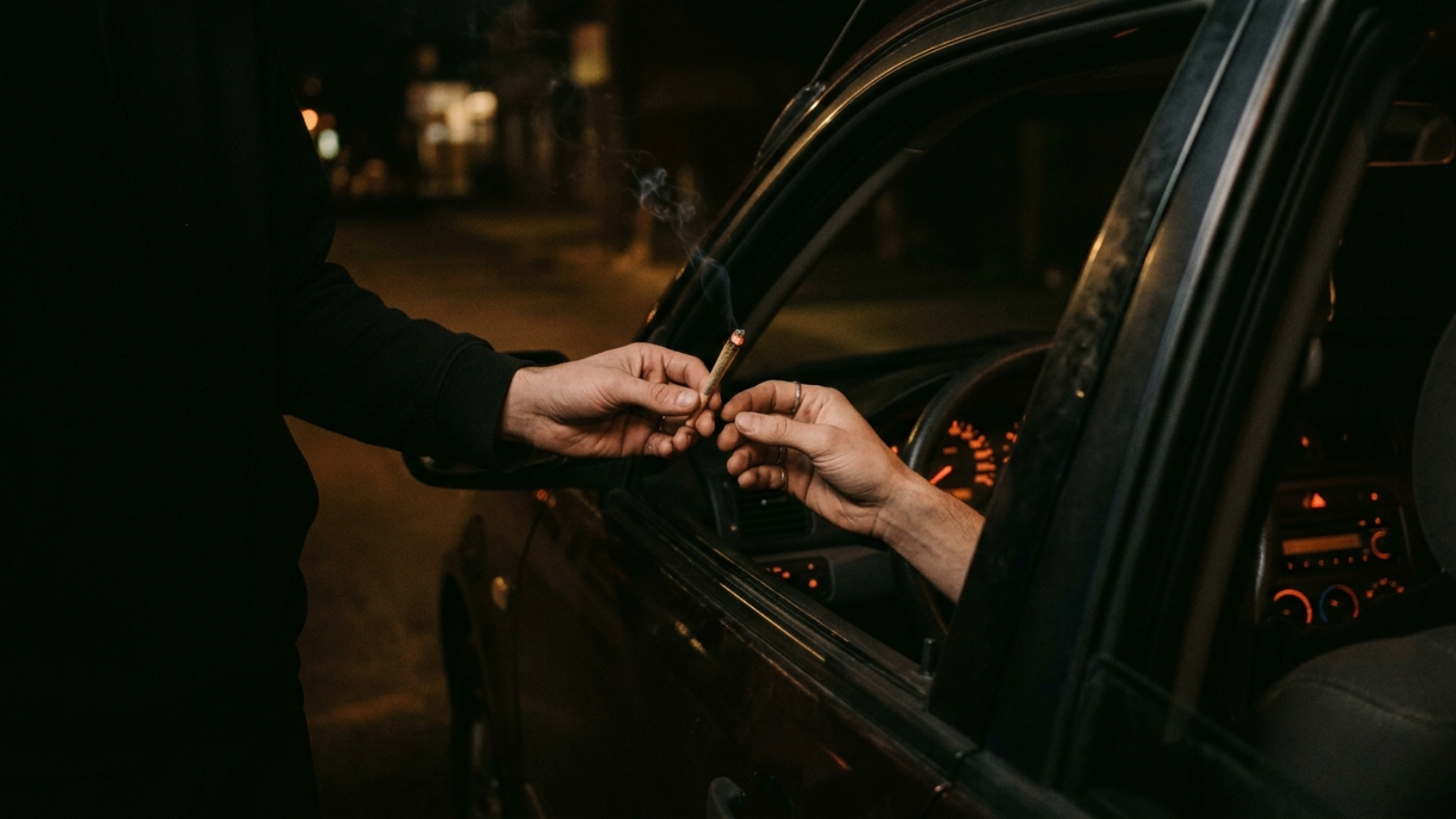 Passing a joint through a car window at night under ember glow lighting