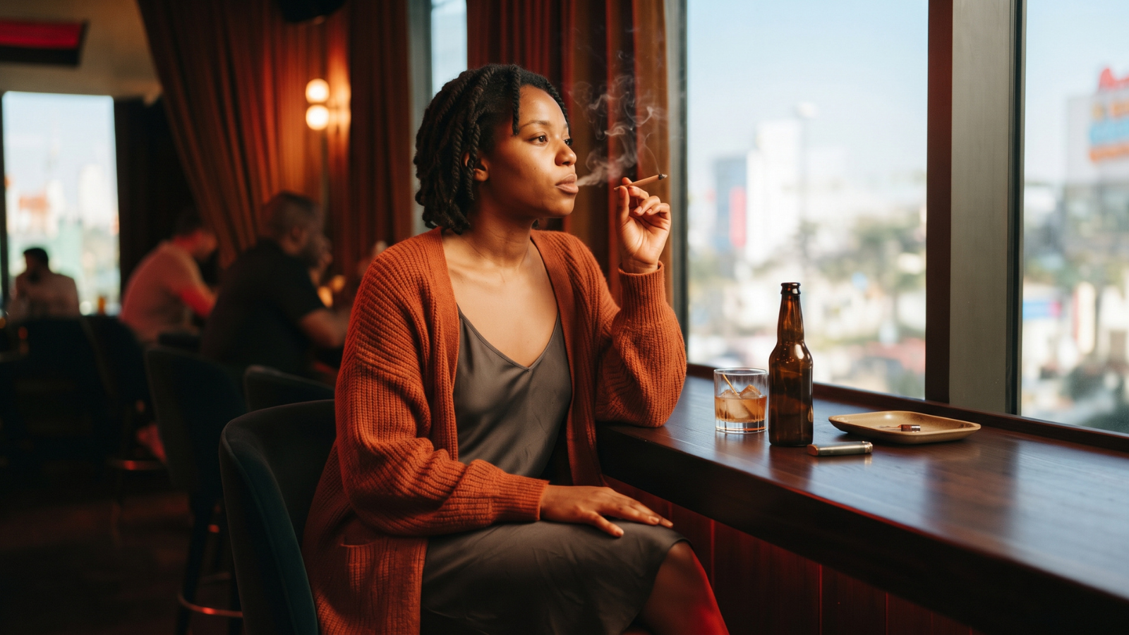 Woman in burnt orange cardigan smoking a joint at a moody hotel bar window.